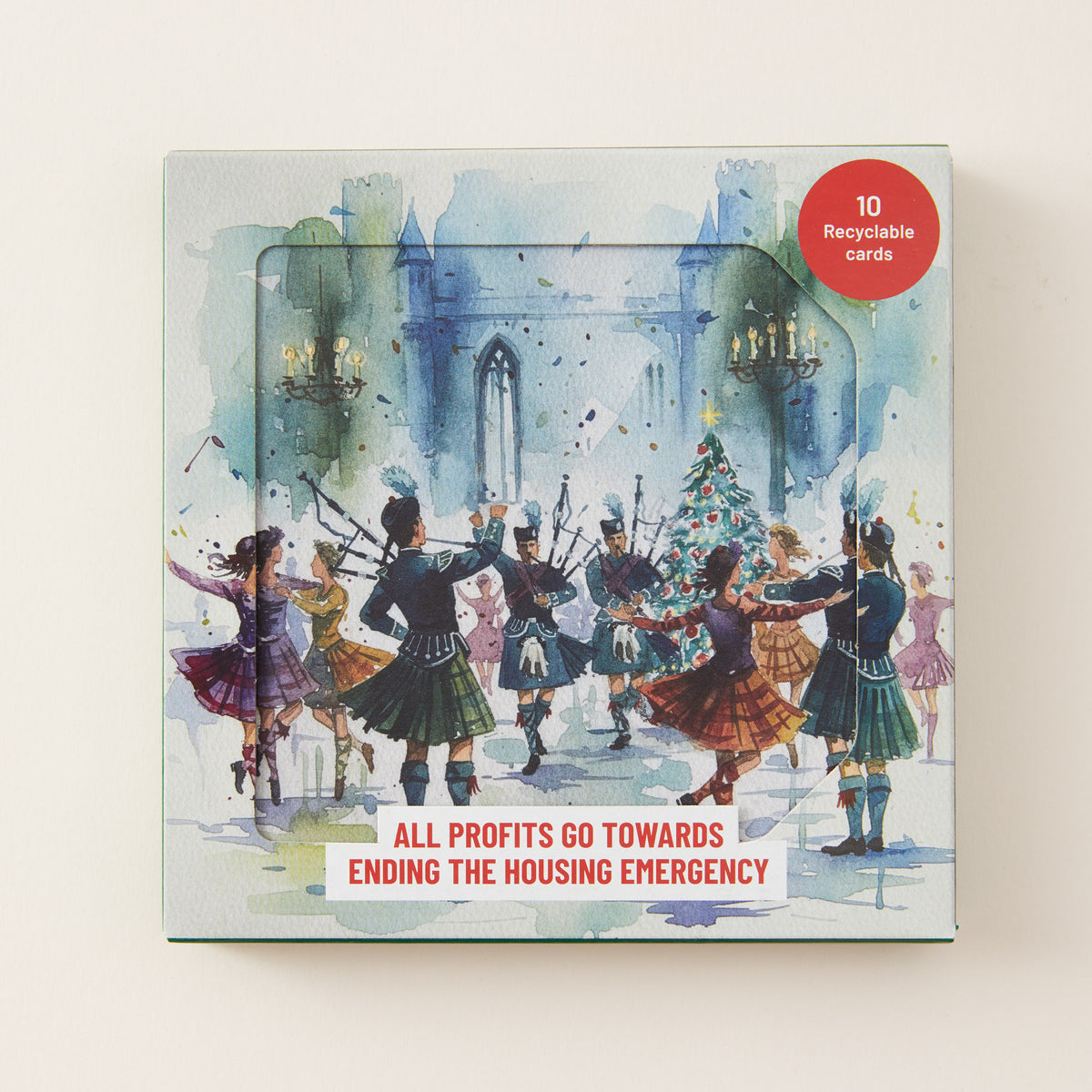 A square printed card deck box with illustrated watercolor-style artwork of people dancing around a Christmas tree in front of a castle. A red circular badge says '10 Recyclable cards,' and a white banner at the bottom reads 'ALL PROFITS GO TOWARDS ENDING THE HOUSING EMERGENCY.