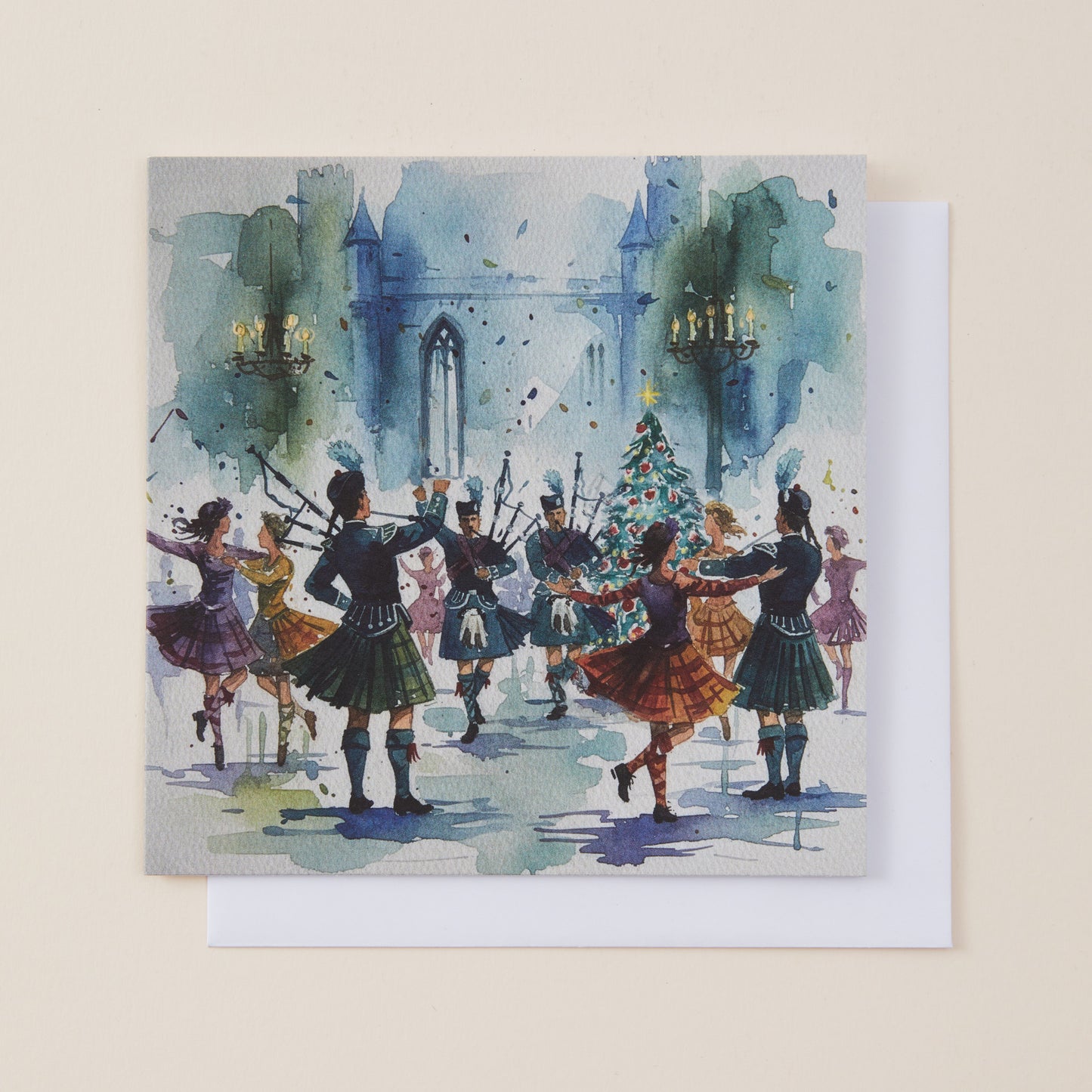 A square art print or greeting card featuring a watercolor illustration of people in Scottish Highland dress dancing around a decorated Christmas tree, with a white envelope behind it on a light beige background.