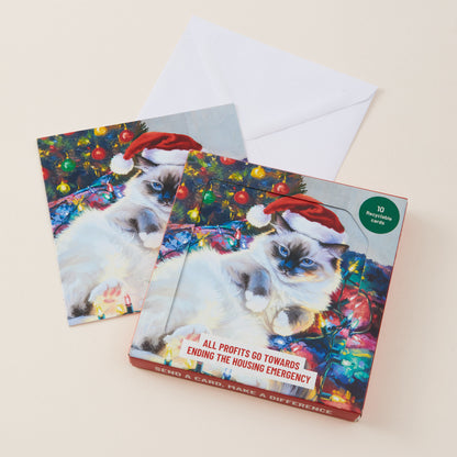 A Christmas-themed card set featuring a fluffy white cat wearing a red Santa hat and hatband, posed in front of a decorated Christmas tree with colorful lights. A white envelope is placed behind the cards on a light beige background.