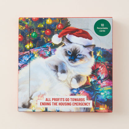 A square product box featuring a colorful illustration of a fluffy white cat wearing a red Santa hat, with Christmas lights and trees in the background. Text on the box reads 'ALL PROFITS GO TOWARDS ENDING THE HOUSING EMERGENCY' and a green circle says '10 Recyclable cards'.