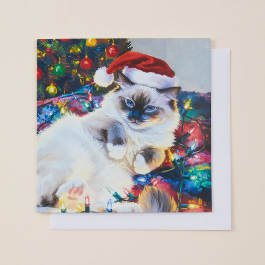 A square Christmas greeting card featuring a painted fluffy cat wearing a Santa hat, surrounded by a decorated Christmas tree and colorful lights, shown with a white envelope on a light beige background.