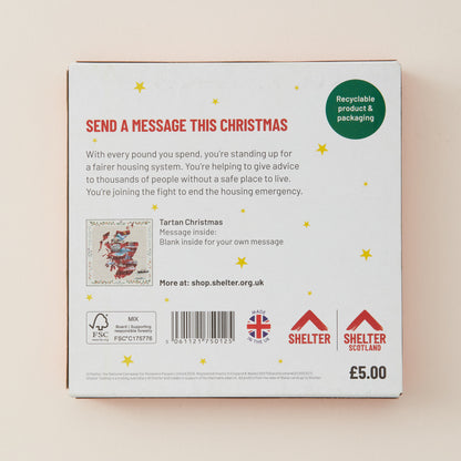 The back of a Tartan Christmas gift box, showing a white surface with red and green text, a message box illustration, a barcode, and Shelter logos, photographed against a light beige background.
