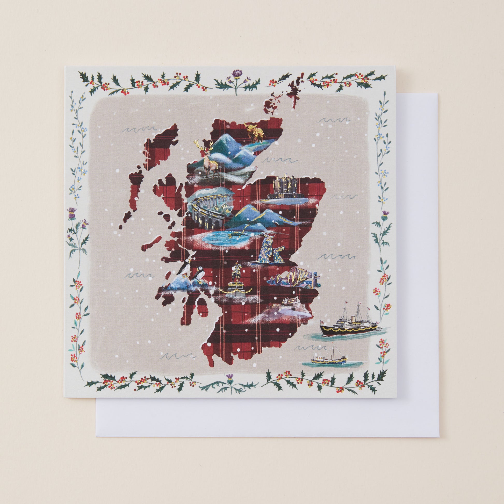 A square greeting card featuring a collage-style map of Scotland in red, blue, and gray tones, surrounded by a decorative holly and floral border, with a white envelope behind it on a light beige background.