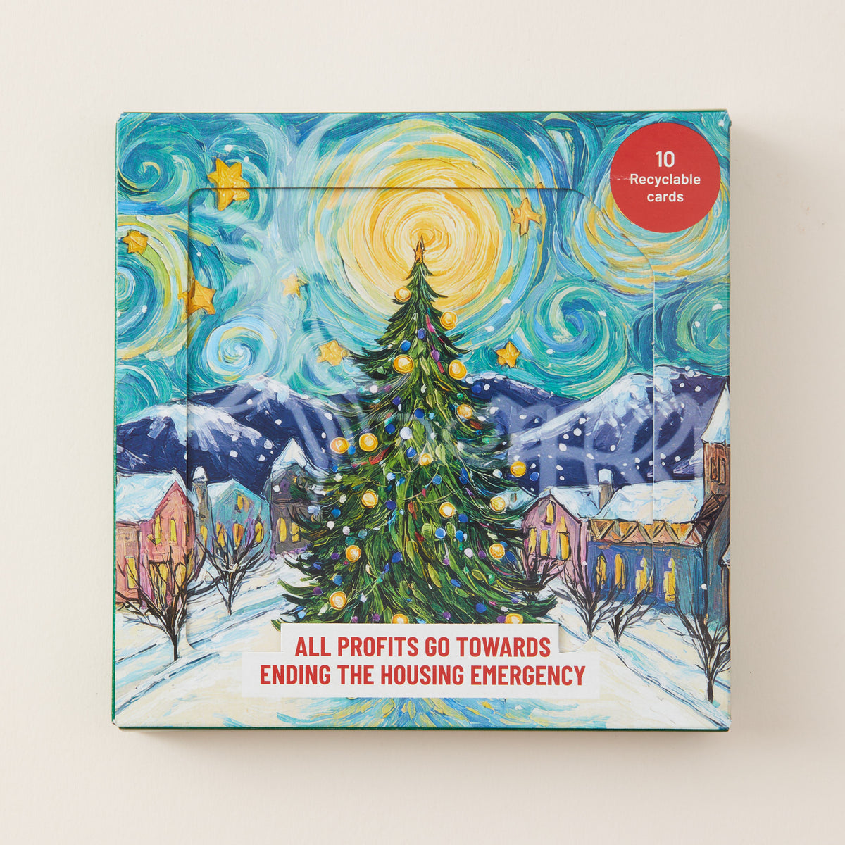 A square printed card set box featuring a stylized snowy village scene with a decorated Christmas tree, mountains, and swirling sky in a starry art style. A red circular badge says '10 Recyclable Cards,' and text at the bottom reads 'ALL PROFITS GO TOWARDS ENDING THE HOUSING EMERGENCY.