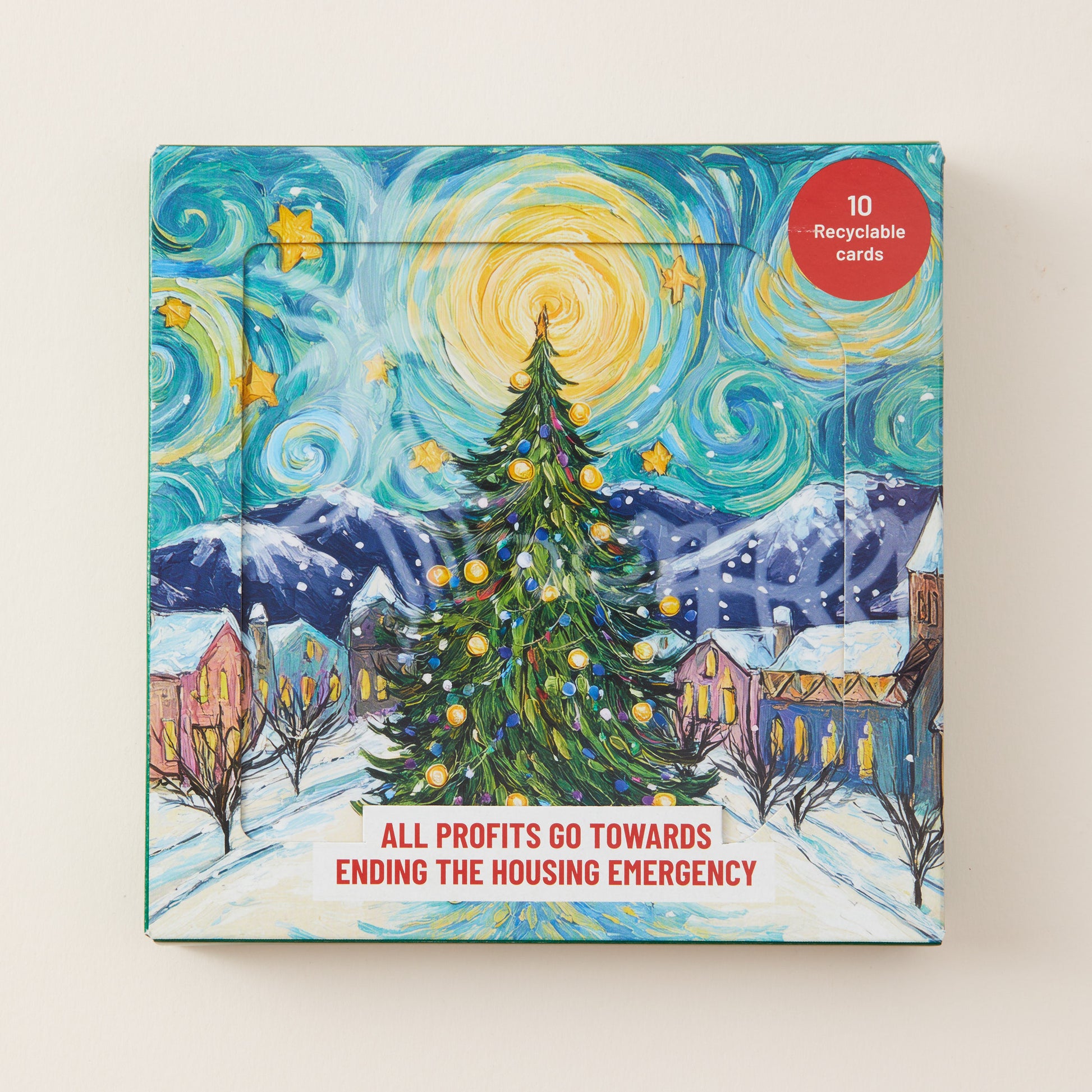 A square printed card set box featuring a stylized snowy village scene with a decorated Christmas tree, mountains, and swirling sky in a starry art style. A red circular badge says '10 Recyclable Cards,' and text at the bottom reads 'ALL PROFITS GO TOWARDS ENDING THE HOUSING EMERGENCY.