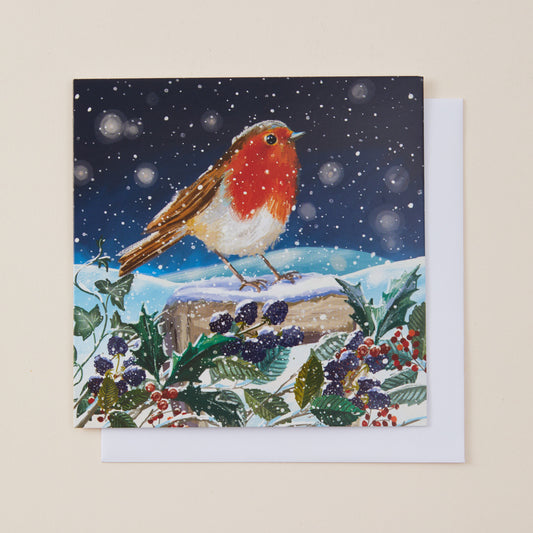 A square greeting card featuring a winter Christmas scene with a robin perched on a snowy log, surrounded by holly and berries under a snowy night sky, shown with a white envelope on a light beige background.