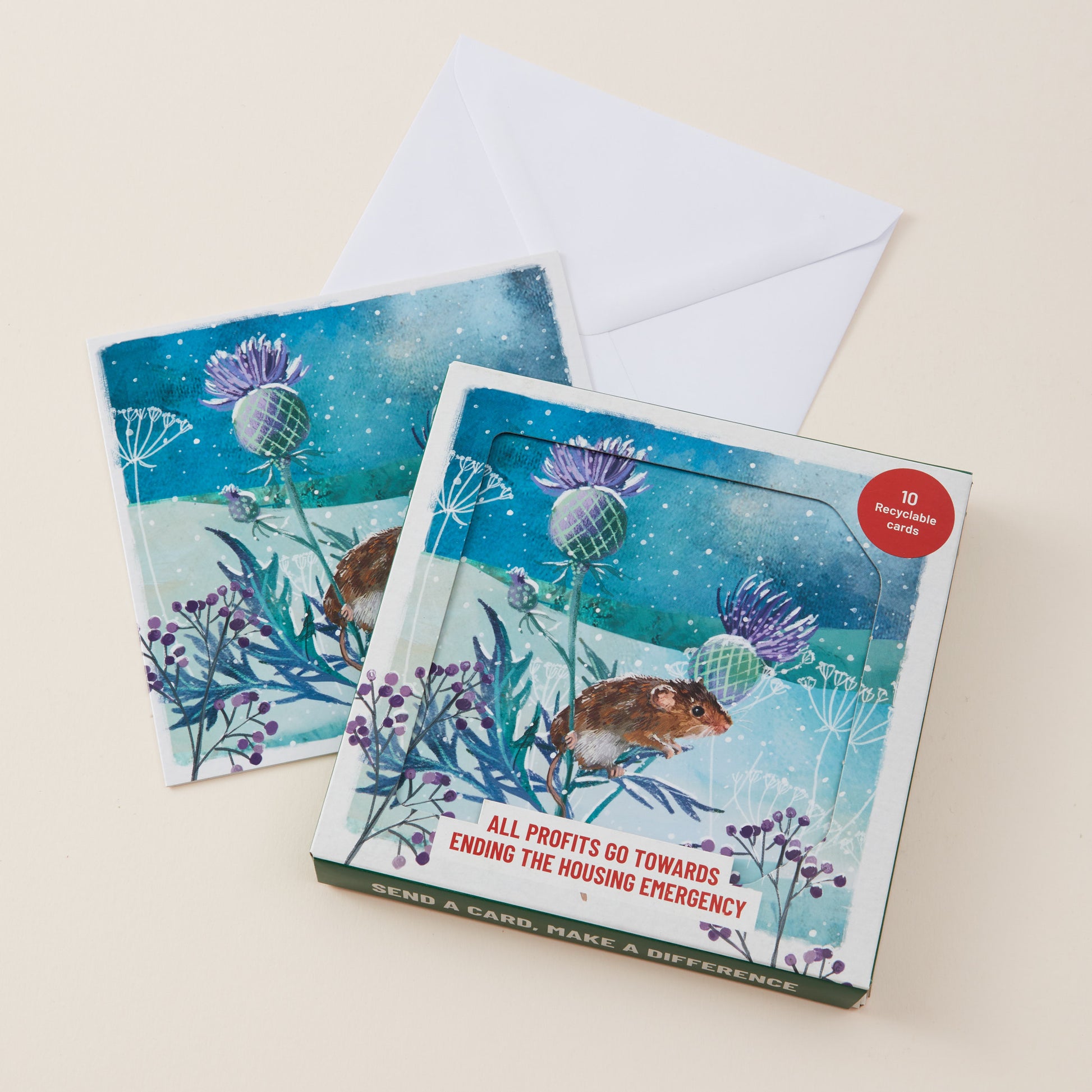 A square greeting card set with an illustrated mouse and purple thistles on a snowy blue background, shown with two cards and a white envelope on a light beige surface. A red circular sticker reads '10 recyclable cards', and the bottom edge says 'SEND A CARD, MAKE A DIFFERENCE'.