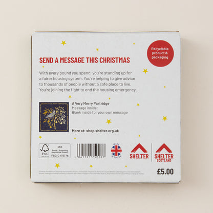 The back of a white cardboard box for Shelter UK, featuring red and black text, a Christmas-themed message, a barcode, and recycling and UK logos, photographed against a light beige background.