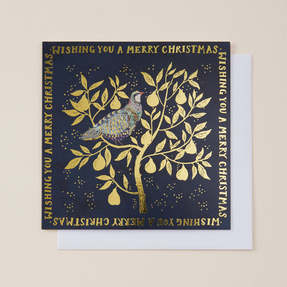 A square Christmas greeting card with a dark navy blue background and metallic gold foil illustration of a bird perched on a tree with pears, shown with a white envelope on a light beige background.