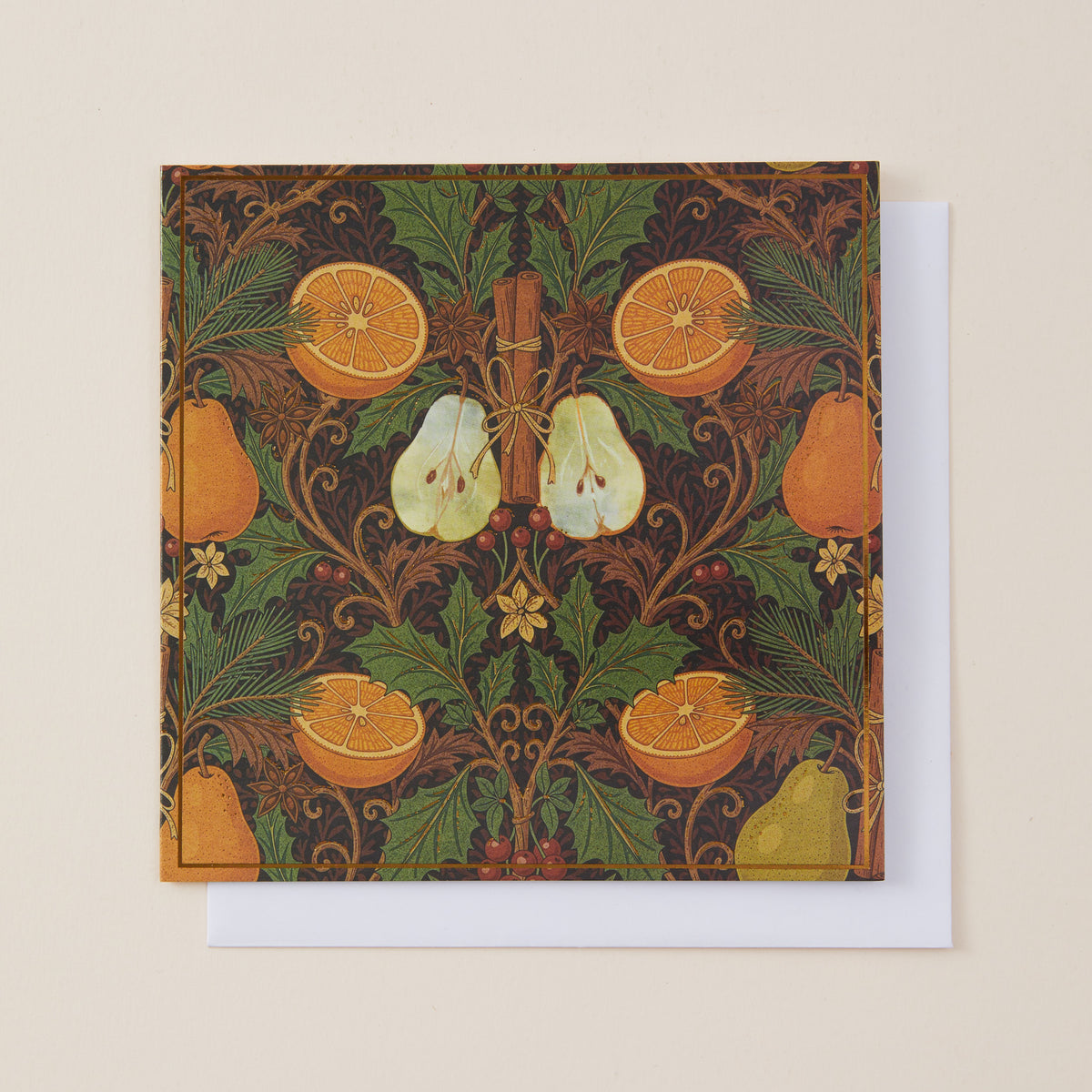 A square art print or card featuring a William Morris-style botanical pattern with oranges, pears, leaves, and a cinnamon stick on a dark background, shown with a white envelope behind it on a light beige surface.