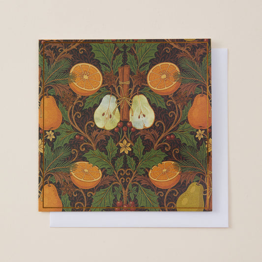 A square art print or card featuring a William Morris-style botanical pattern with oranges, pears, leaves, and a cinnamon stick on a dark background, shown with a white envelope behind it on a light beige surface.