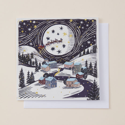 A square Christmas greeting card featuring an illustrated winter village scene under a large moon and starry night sky, with Santa Claus in a sleigh. The card is shown with a white envelope behind it on a light beige background.