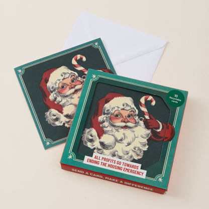A set of Christmas greeting cards and envelopes shown on a light beige background. The front card features a vintage-style illustration of Santa Claus with the text 'ALL PROFITS GO TOWARDS ENDING THE HOUSING EMERGENCY' and 'SEND A CARD, MAKE A DIFFERENCE' on the side.