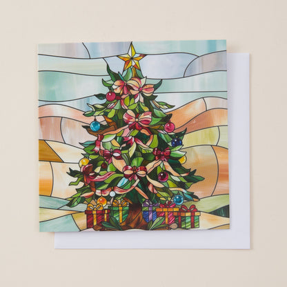 A square Christmas greeting card with a stained-glass style illustration of a decorated tree, shown with a white envelope on a light beige background.