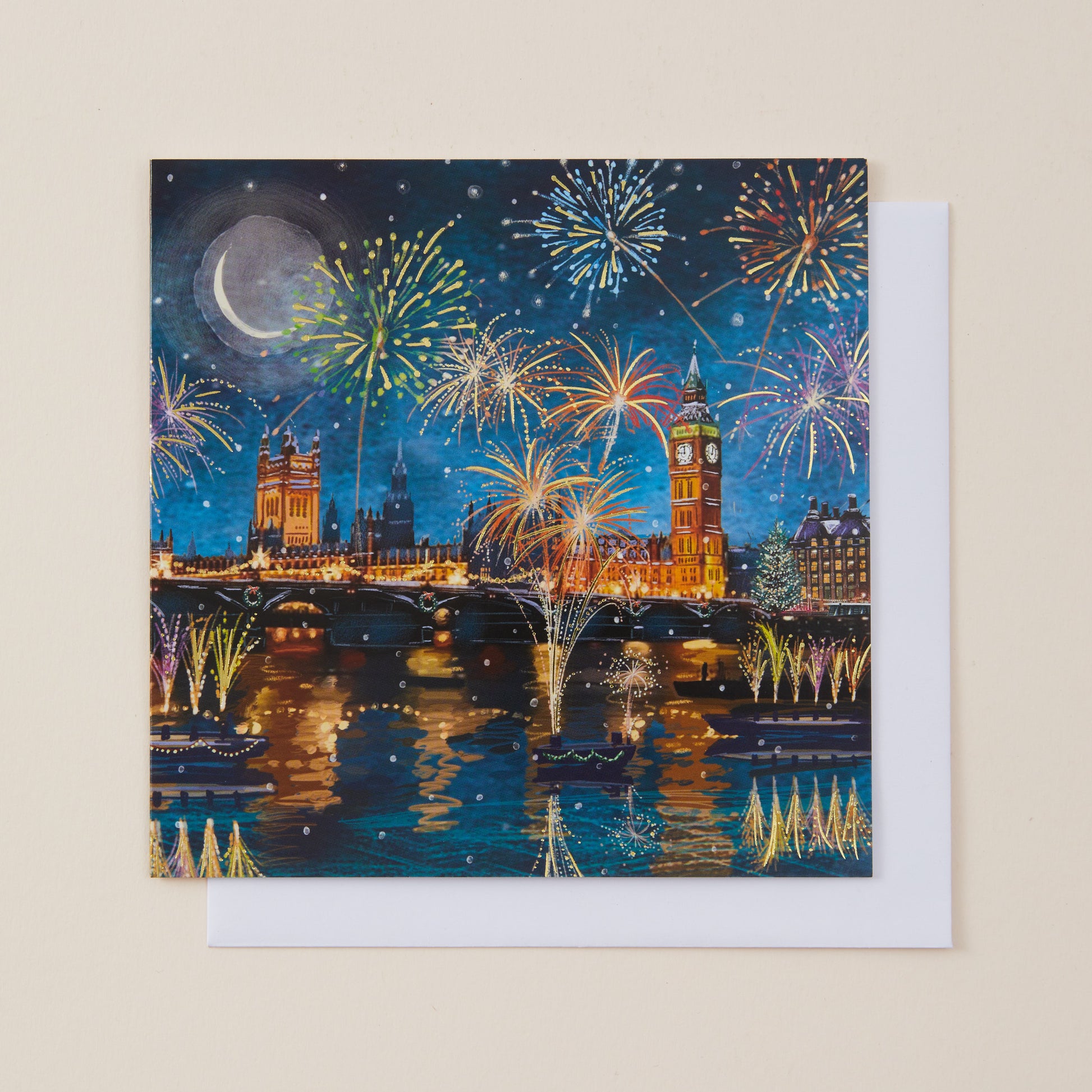 A square Christmas card featuring a colorful night scene of London with fireworks, Big Ben, Westminster Bridge, and reflections on the Thames, shown with a white envelope behind it on a light beige background.
