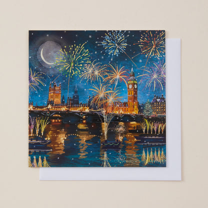 A square Christmas card featuring a colorful night scene of London with fireworks, Big Ben, Westminster Bridge, and reflections on the Thames, shown with a white envelope behind it on a light beige background.