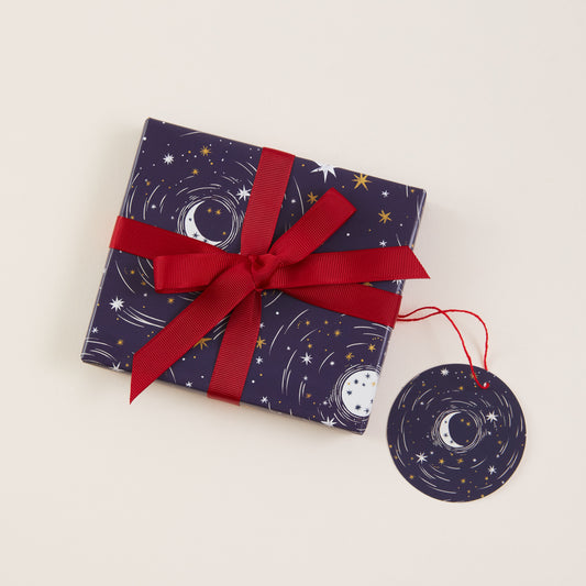 A square gift box wrapped in dark navy paper with a celestial print of stars and crescent moons, tied with a wide red ribbon and accompanied by a matching round tag on a light beige background.
