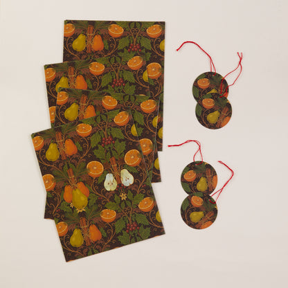A flat-lay of decorative paper goods featuring a William Morris-style botanical pattern with oranges, pears, and berries on a dark background, alongside two round hanging tags with red strings, all arranged on a light cream surface.