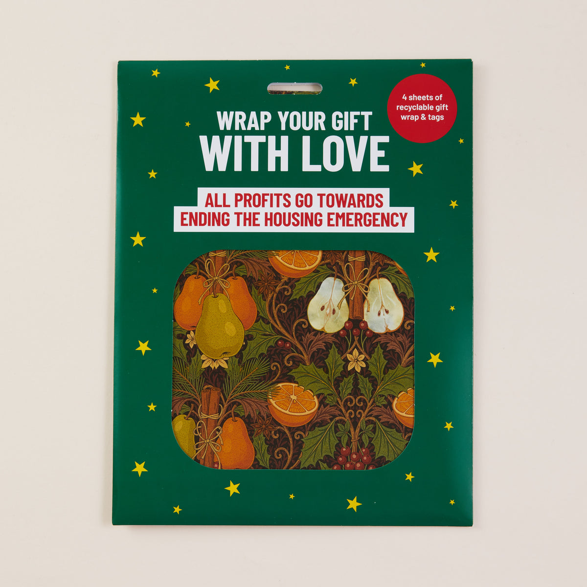 A packaged gift wrap set on a dark green card with yellow stars and a central cutout showing fruit-patterned wrap sheets. The package text reads 'WRAP YOUR GIFT WITH LOVE' and 'ALL PROFITS GO TOWARDS ENDING THE HOUSING EMERGENCY.