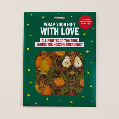 A packaged gift wrap set on a dark green card with yellow stars and a central cutout showing fruit-patterned wrap sheets. The package text reads 'WRAP YOUR GIFT WITH LOVE' and 'ALL PROFITS GO TOWARDS ENDING THE HOUSING EMERGENCY.