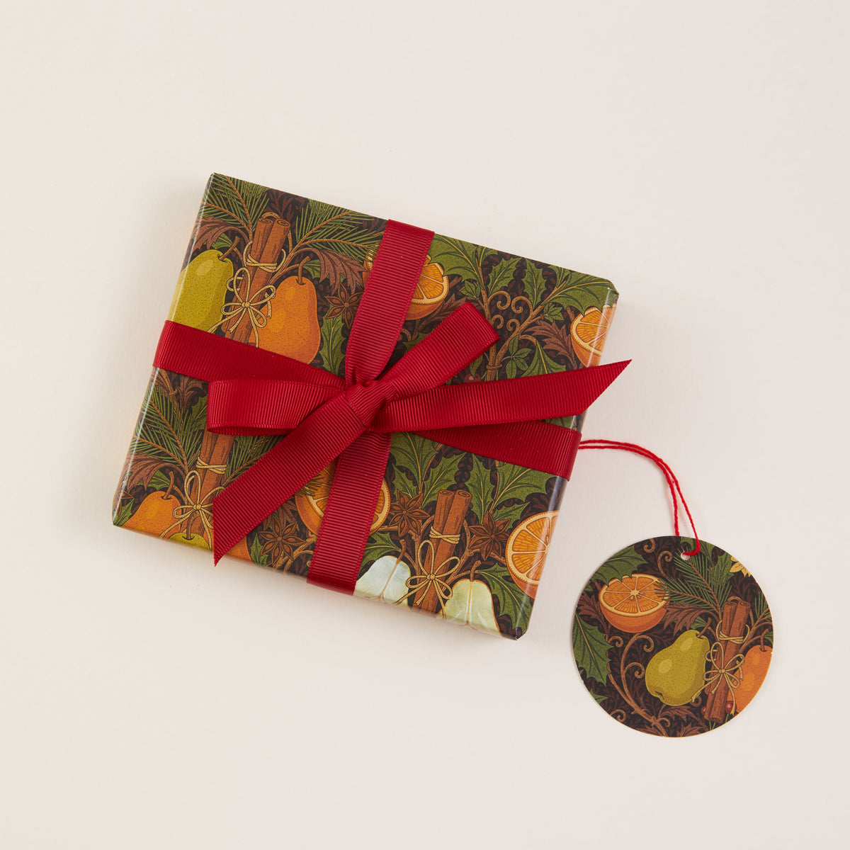 A small gift box wrapped in a dark botanical and fruit patterned paper with a red ribbon bow and a matching round hanging tag, photographed on a plain light background.