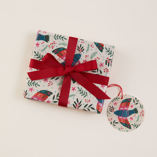 A gift box wrapped in white paper with a colorful bird and floral pattern, tied with a dark red ribbon and bow, with a matching round tag hanging from the ribbon, on a light beige background.