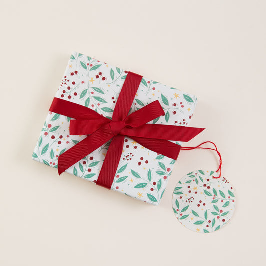 A gift box wrapped in white paper with a green and red berry leaf pattern, tied with a wide red ribbon and bow, on a light beige background with a matching round tag.