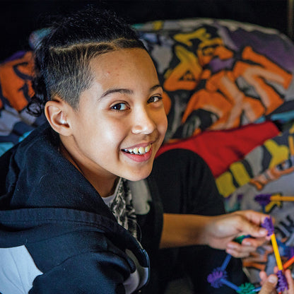 A smiling young person with short dark hair sits on a bed, holding a colorful plastic toy. The room has graffiti-patterned bedding, and a black nightstand is visible in the background.