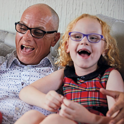 A smiling bald man wearing black-rimmed glasses sits on a couch with a young girl laughing. The man wears a light patterned button-up shirt, and the girl wears a red plaid dress.