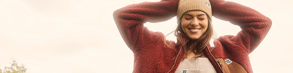 Model smiling with her hands on her  head. She is wearing a burgundy Lucy & Yak fleece and beige dungarees and beanie hat.