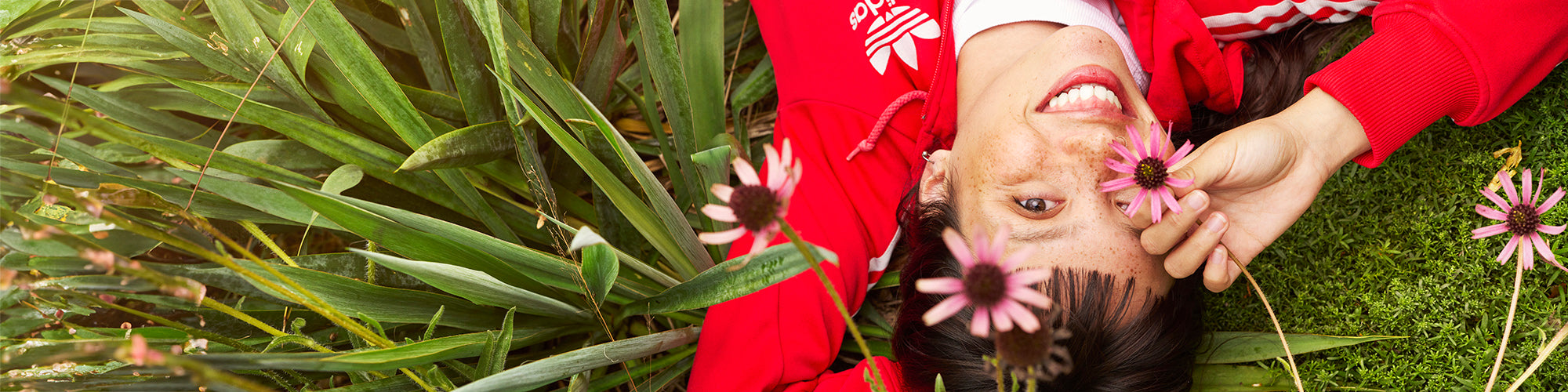 Model lying down in long grass holding a flower over one eye. She is wearing a red tracksuit top.