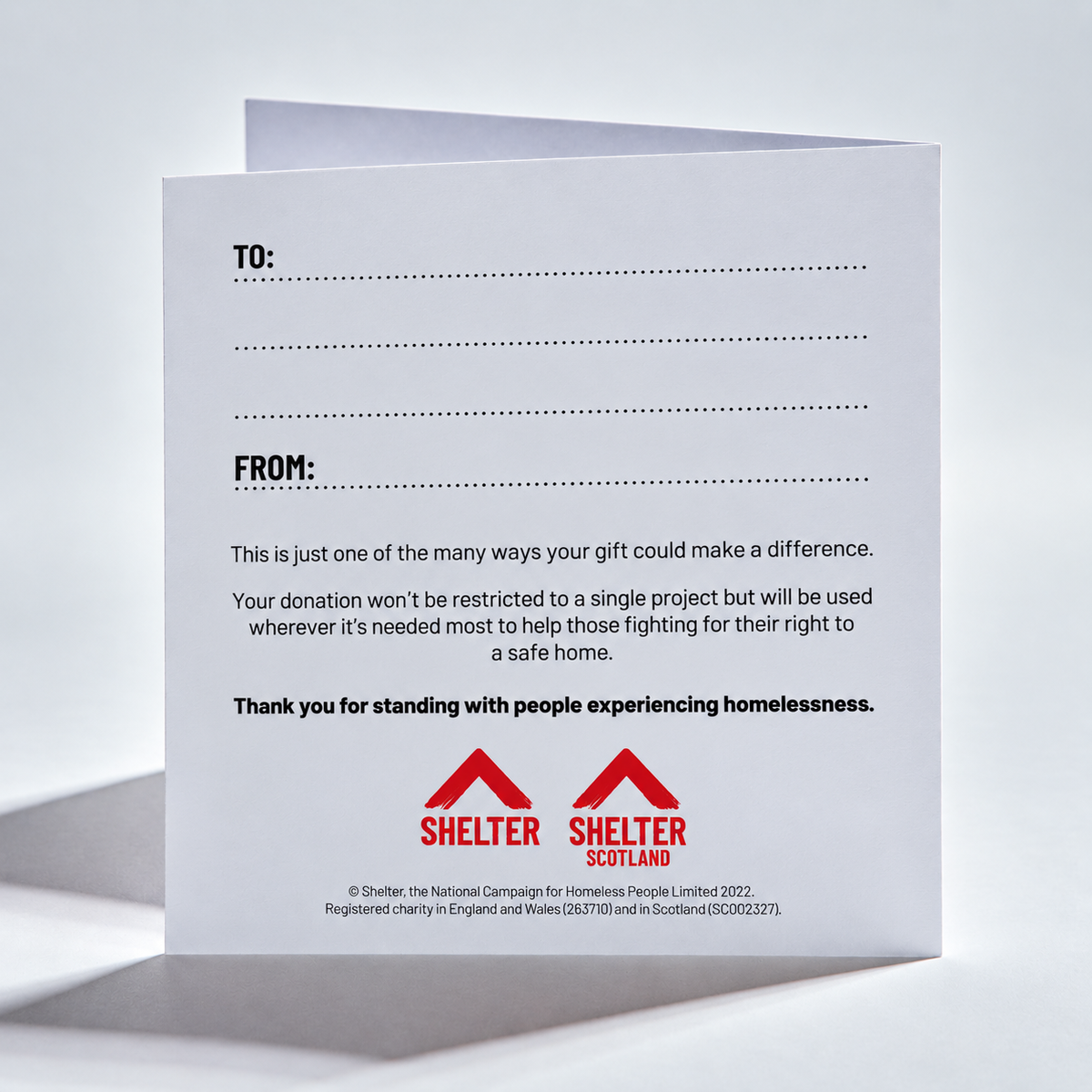A white printed card or envelope shown standing open against a light gray studio background. The front has dotted lines for TO and FROM fields, a paragraph of text about donations helping fight homelessness, and two red Shelter Scotland logos at the bottom.