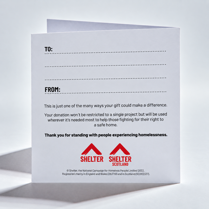 A white printed card or envelope shown standing open against a light gray studio background. The front has dotted lines for TO and FROM fields, a paragraph of text about donations helping fight homelessness, and two red Shelter Scotland logos at the bottom.