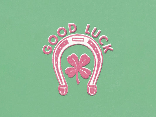 A pink and white horseshoe and clover design against a green background, with pink text: 'GOOD LUCK'.