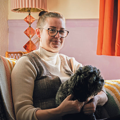 A woman with glasses and her hair in a bun sits on a couch in a cozy room, holding a small black dog. She is wearing a cream turtleneck sweater and a brown knit vest.