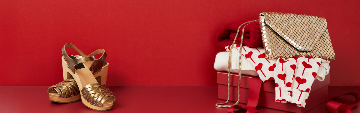 A styled fashion arrangement on a red background featuring a pair of metallic gold strappy sandals on the left and a matching metallic gold woven clutch bag on the right, displayed with a red gift box and a white garment printed with red hearts.