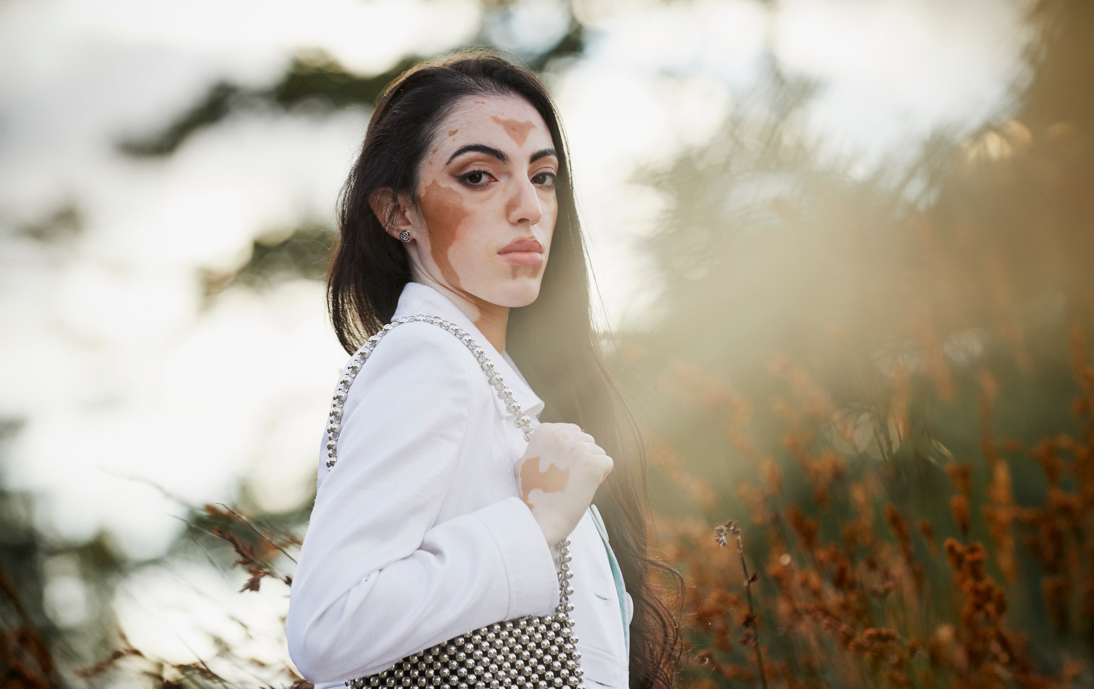 Model standing in autumnal outdoor setting. She wears a white jacket and silver bag on one shoulder.