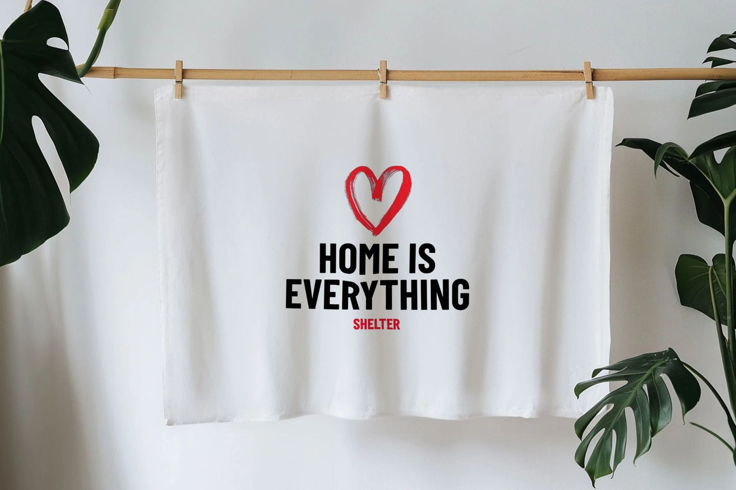 White tea towel hanging pegged up. The teatowel is printed with the phrase 'Home is Everything' in black text under a red heart.