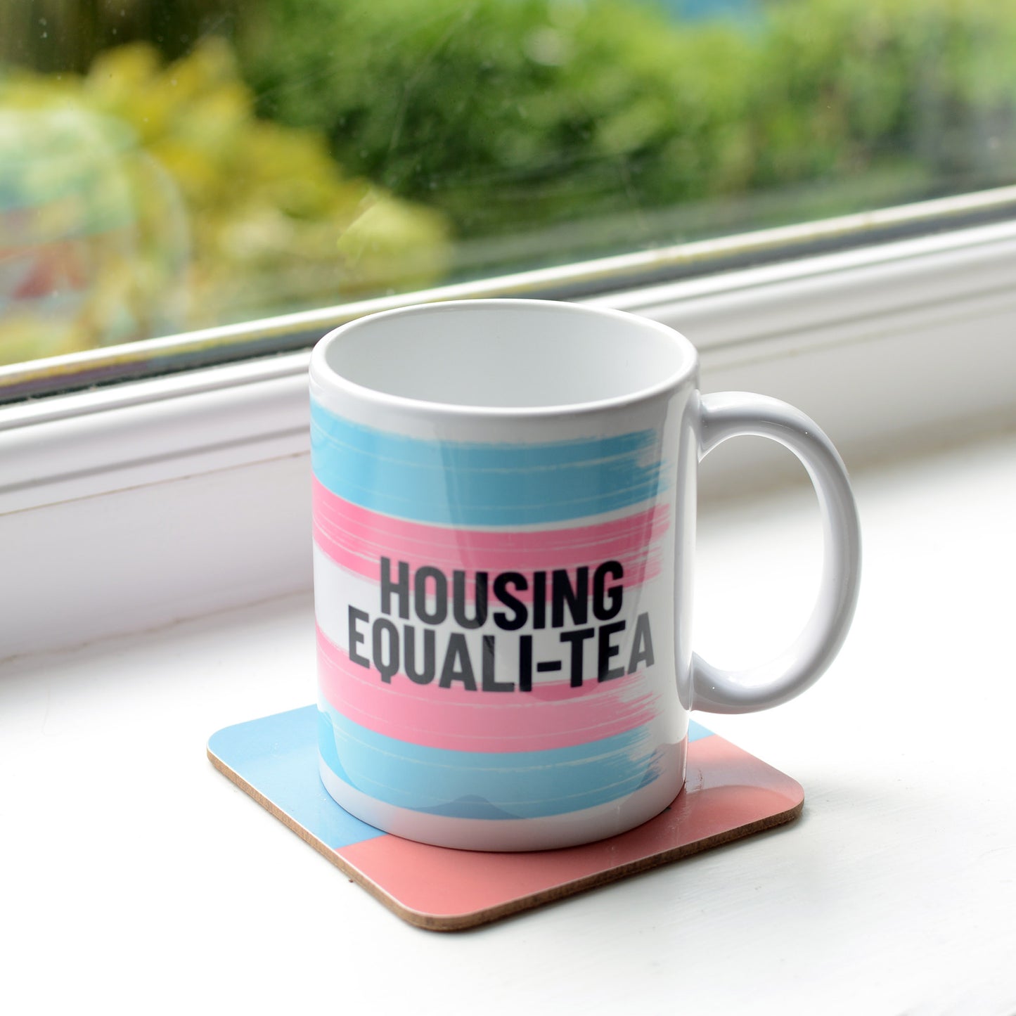 White Housing Equali-Tea Mug with blue and pink background represeting the trans flag. The mug is sitting on a window sill. 