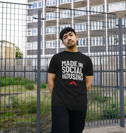 Model is wearing a black T-shirt with the slogan 'Made in social housing' in bold, white text. He is standing in front of a metal fence with a grassy area and a block of flats behind. 
