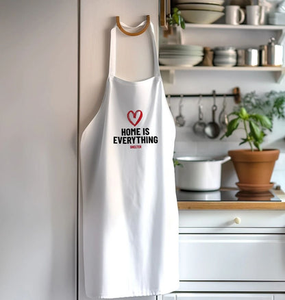 A white apron hanging up in a kitchen. The apron has a red heart design and the wording 'HOME IS EVERYTHING'