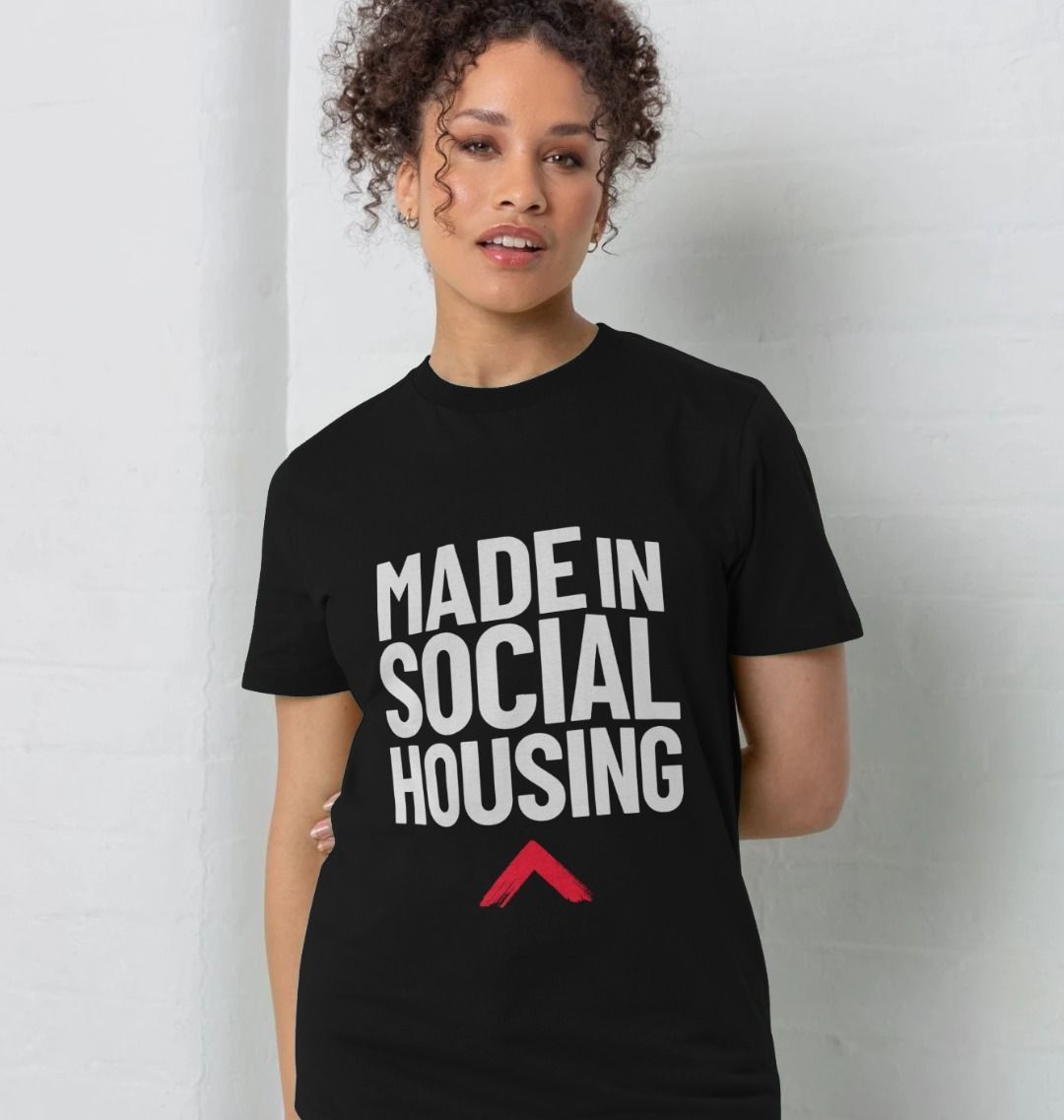 Model is standing with one arm behind her back. She is wearing a black T-shirt with the slogan 'Made in social housing' in bold, white text.