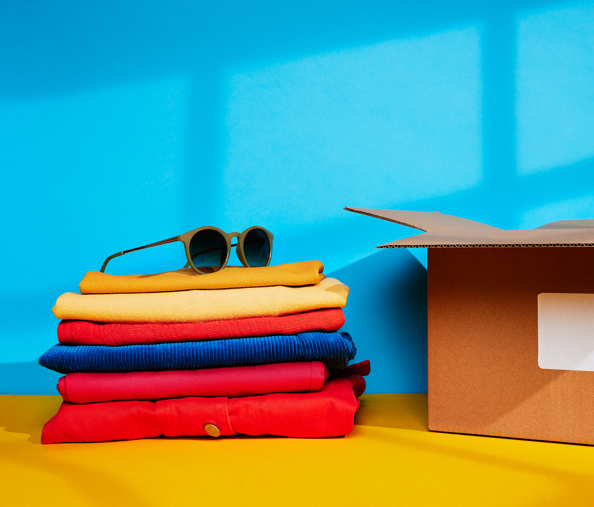 Stack of colorful folded clothes with sunglasses on top, next to a cardboard box against a blue wall.