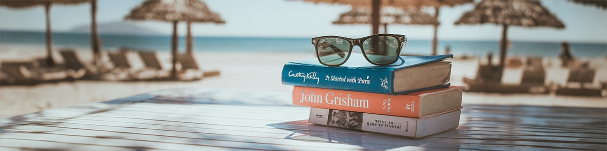 a pile of paperback books on a table with sunglasses on top. Blurred but visible in the background is a beach scene with sun loungers