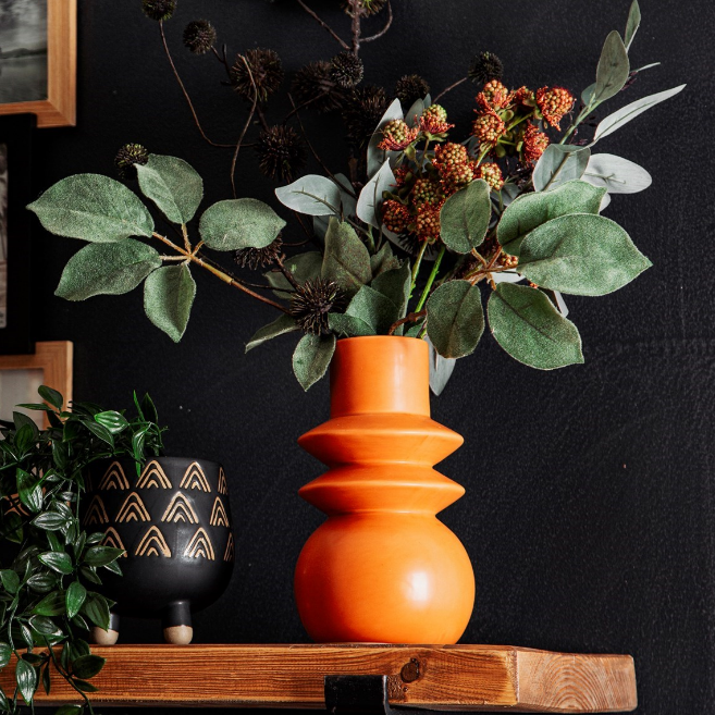 terracotta vase on a teak shelf against a black painted wall with dried flowers inside the vase