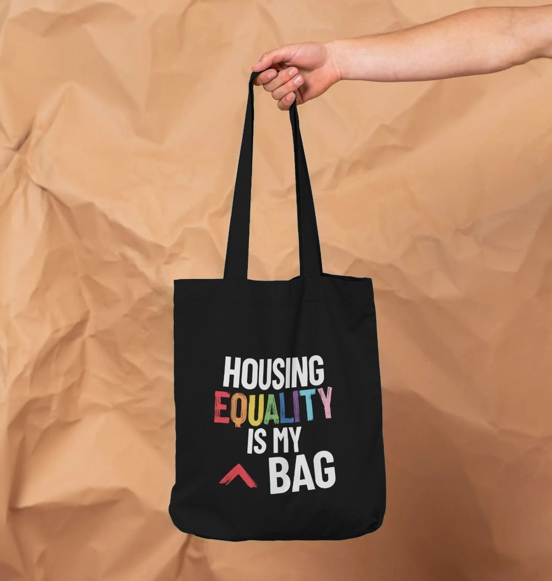 black tote bag design, with slogan in white bold text, the word equality is in the LGBTQ+ flag colours, bag reads "Housing equality is my bag"